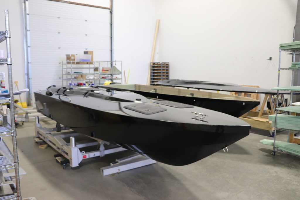 Two black boat hulls are positioned on stands in a workshop with shelves, tools, and materials visible in the background.
