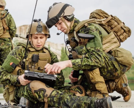 Two soldiers in camouflage uniforms and tactical gear use a rugged tablet outdoors; one is sitting, the other is crouching and pointing at the device.