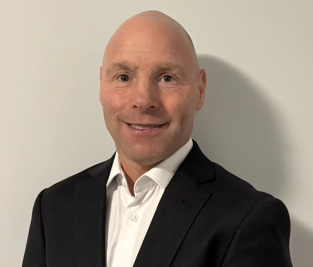 A bald man in a dark suit jacket and white shirt stands in front of a plain light-colored wall, smiling at the camera.