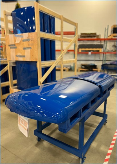 A shiny blue industrial part rests on a wheeled metal cart in a warehouse, with wooden crates and shelves visible in the background.