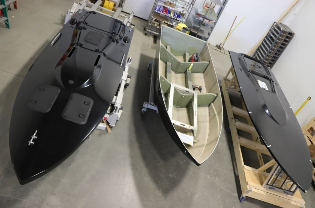 Three boat hulls at different stages of construction are arranged side by side in a workshop, with tools and materials visible in the background.