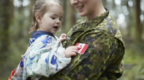 A child in patterned clothing touches a Canadian flag patch on the uniform of a smiling person in military camouflage, outdoors in a wooded area.