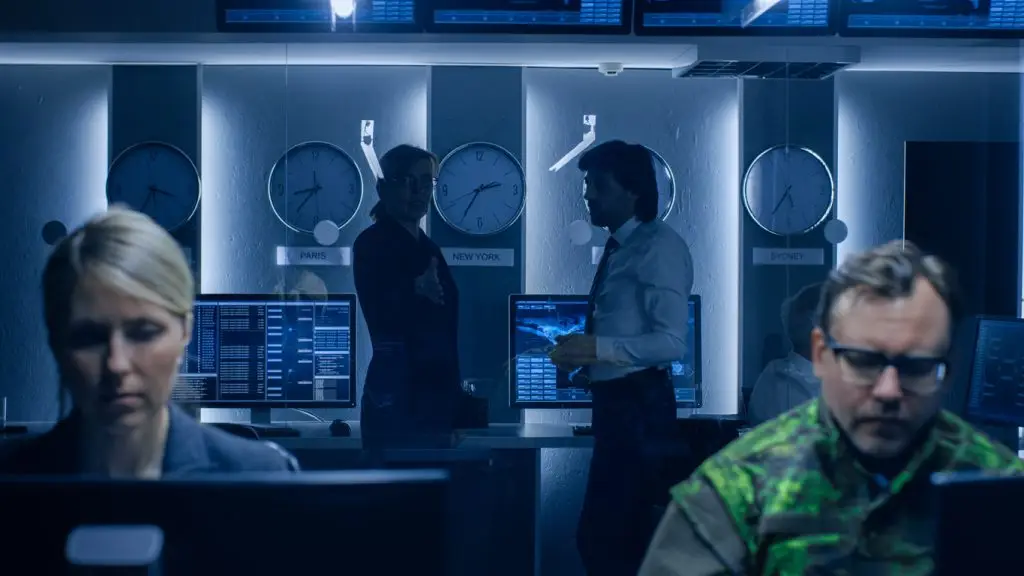 People work at computer stations in a dimly lit control room with wall clocks showing different time zones and large monitors displaying data.