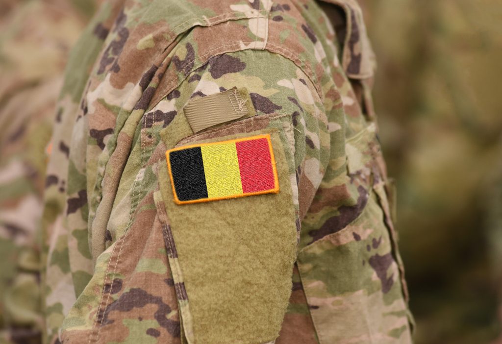 A close-up of a person wearing camouflage military uniform with a Belgian flag patch on the sleeve.