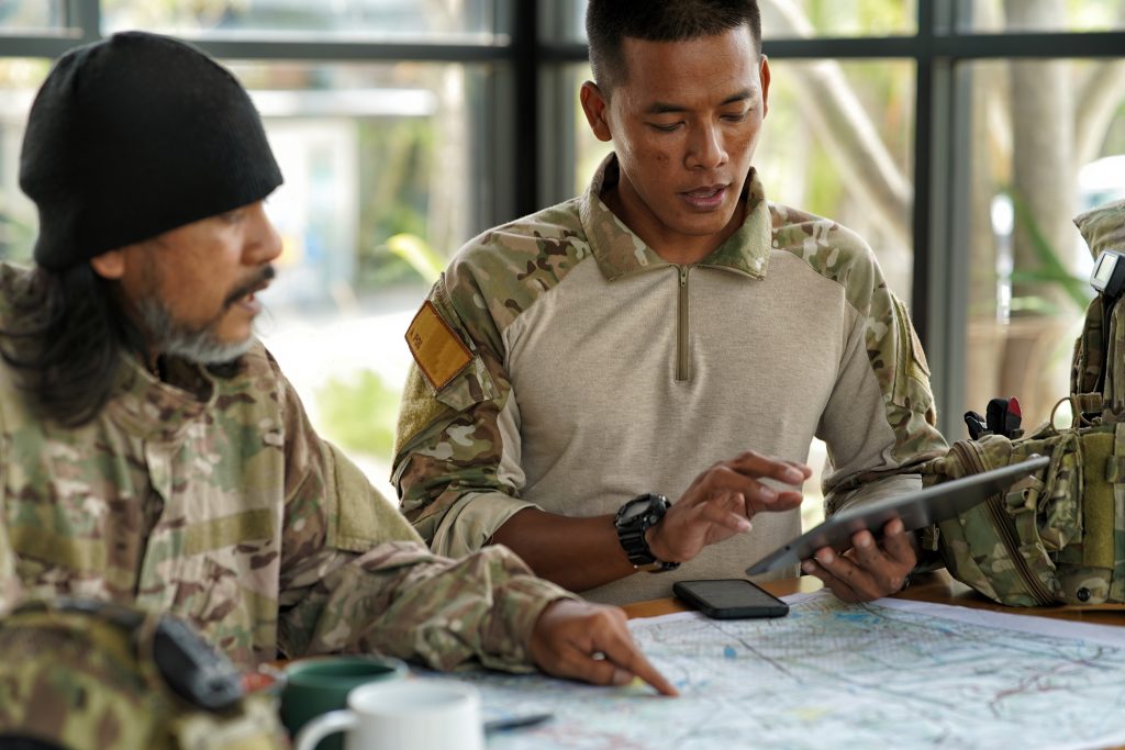 Two men in military camouflage uniforms review a map on a table; one uses a tablet while the other points at the map.