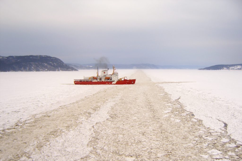 A red icebreaker ship moves through a frozen, ice-covered body of water, leaving a clear path behind it under a cloudy sky with snow-covered hills in the distance.