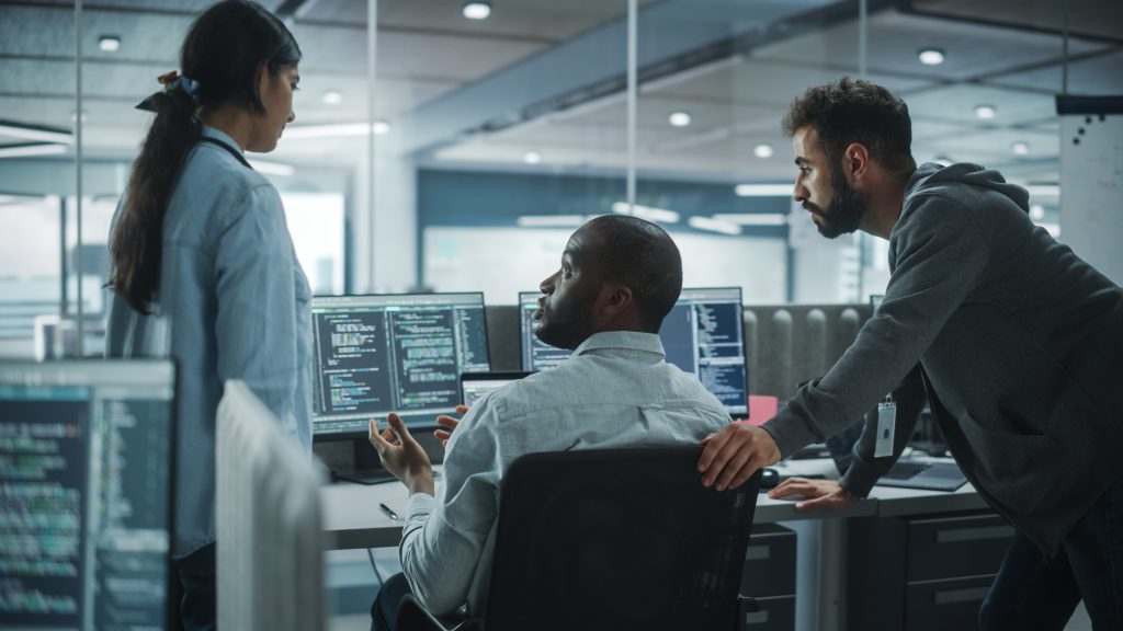 Three people discuss code displayed on computer monitors in a modern office, with one person seated and two standing nearby, all focused on the screens.