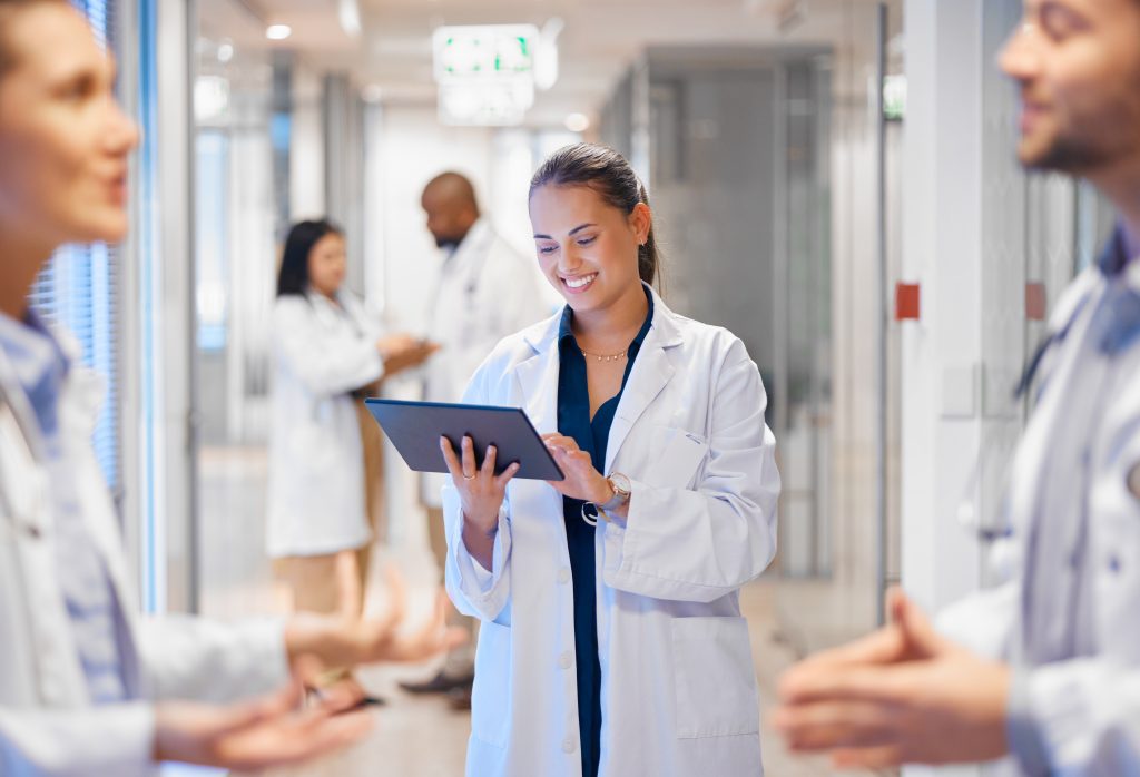 A female doctor in a white coat reviews information on a tablet in a hospital corridor, while other medical professionals converse in the background.