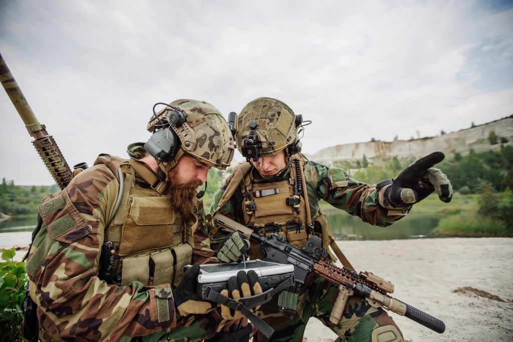 Two soldiers in camouflage uniforms and helmets examine a map or device outdoors, with one pointing into the distance.
