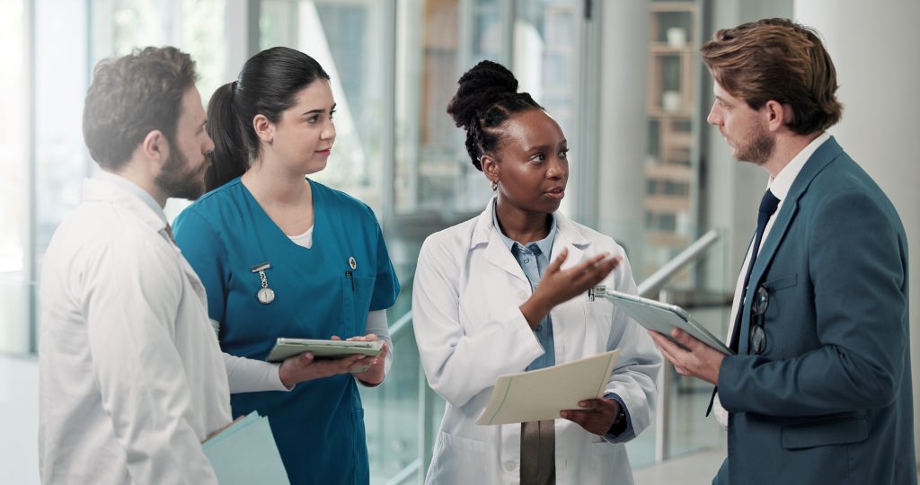 Four medical professionals, two in white coats and two in scrubs, stand together indoors, discussing something while holding tablets and documents.