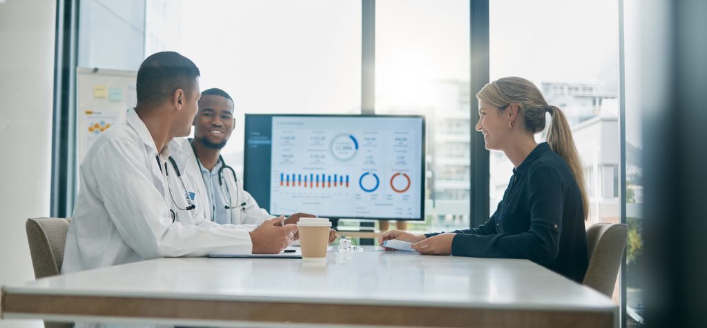 Three people, including two doctors and a woman, sit at a table discussing medical data displayed on a screen in a modern office setting.
