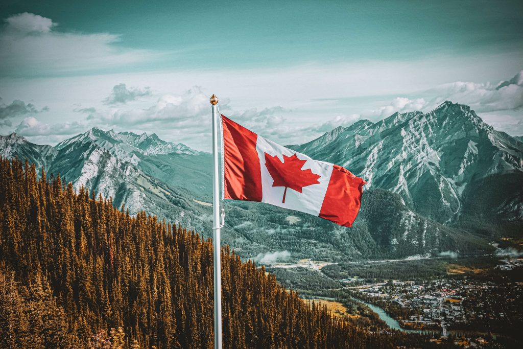 A Canadian flag on a pole stands in front of a forested mountain range and a distant valley under a partly cloudy sky.