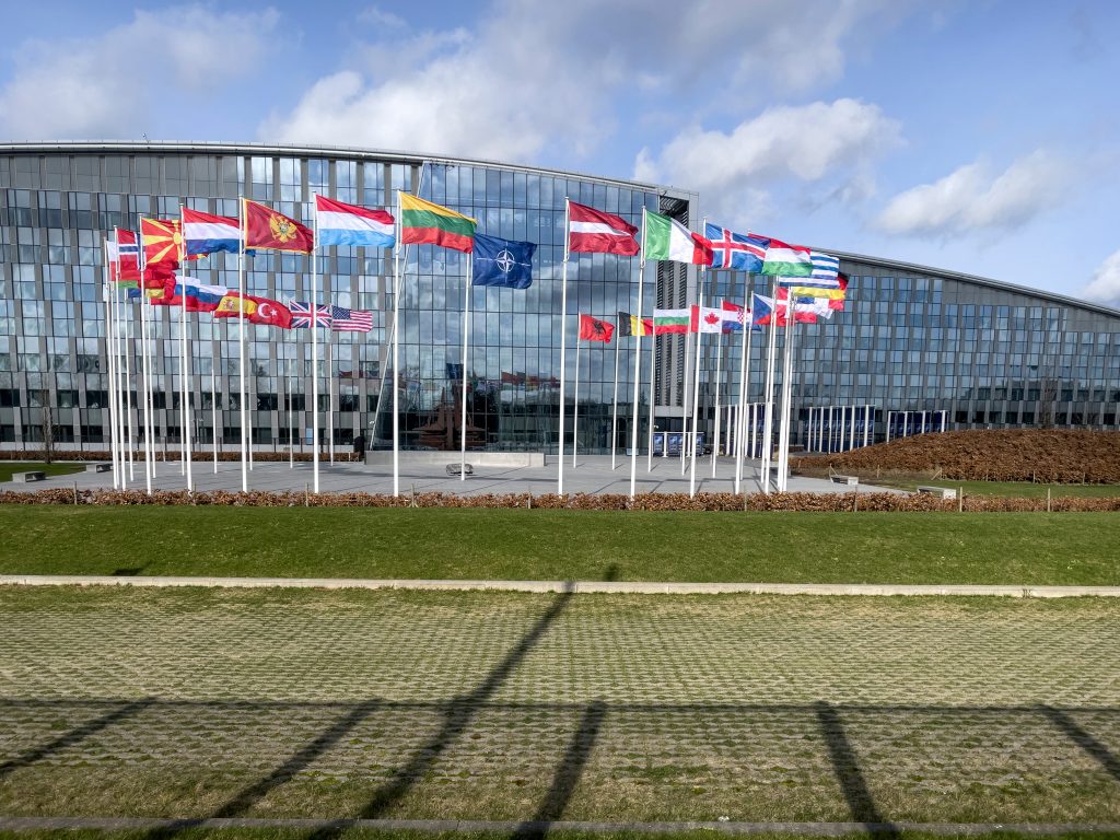 A row of national flags and the NATO flag stand in front of a modern glass building on a sunny day.