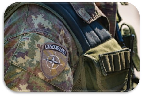 Close-up of a soldier's uniform with a NATO-OTAN patch and tactical vest, showing camouflage pattern and military gear.