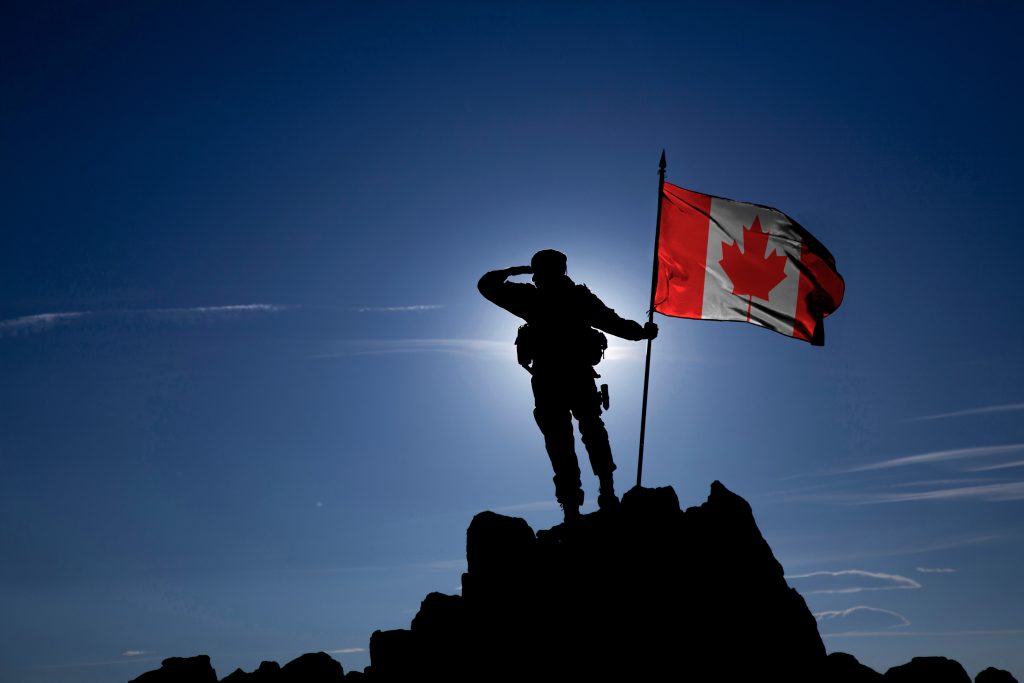 Silhouette of a soldier saluting while holding a Canadian flag on a rocky peak, with a bright sky and sun in the background.