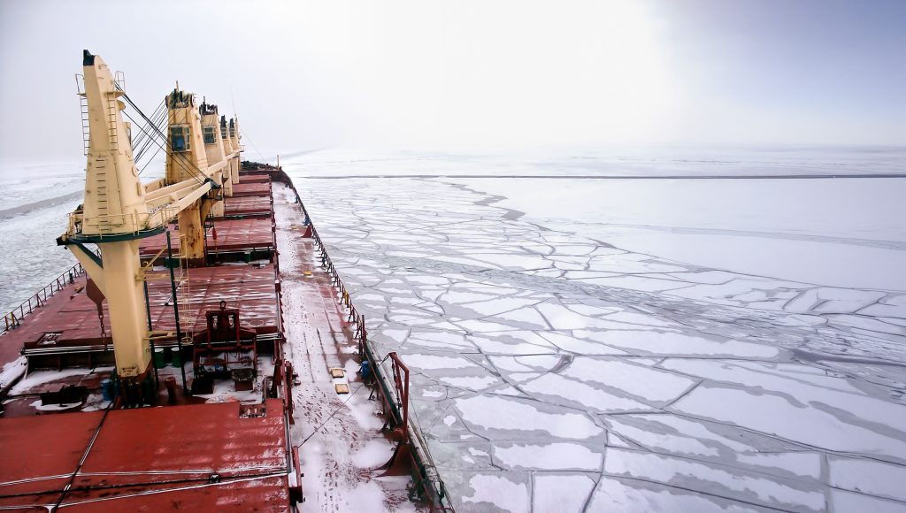 Large cargo ship travels through icy water, breaking up sheets of ice under a cloudy sky.