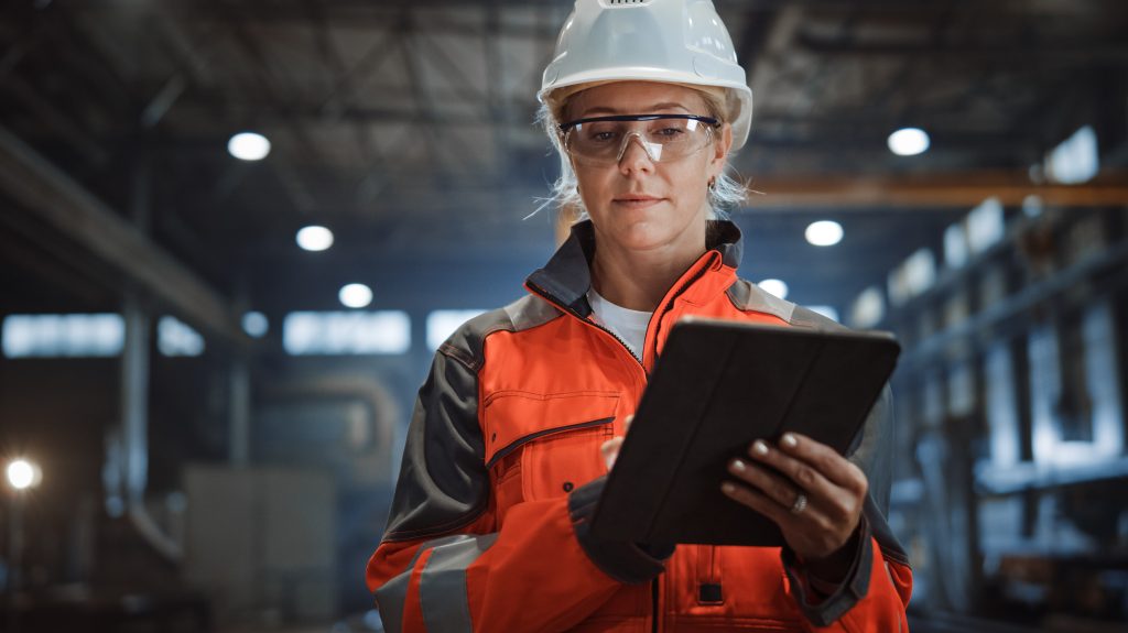 A person in safety gear, including a hard hat and protective glasses, using a tablet in an industrial setting.