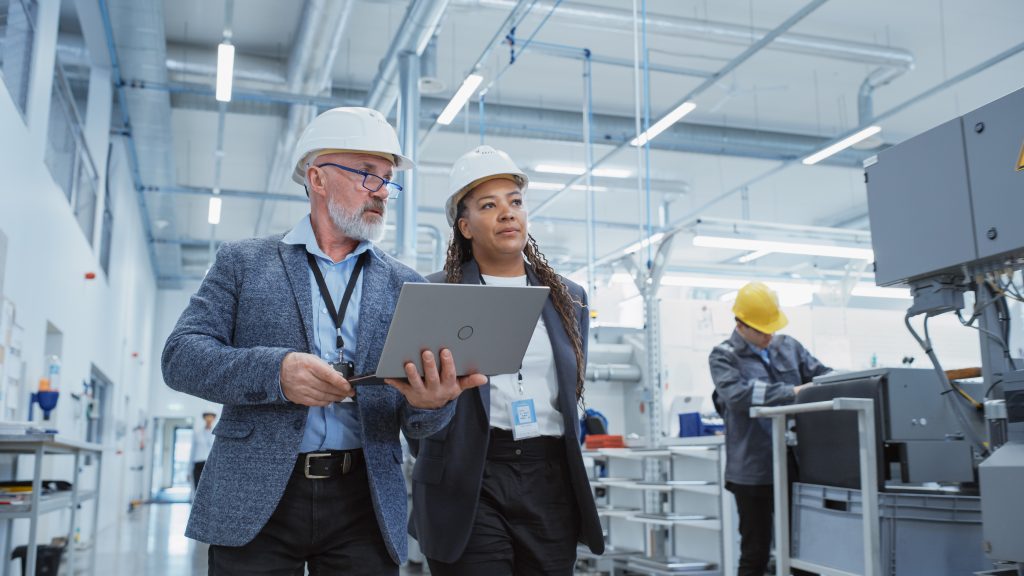 Two people in safety helmets and formal attire discussing something on a laptop in an industrial setting. Another worker, also in a helmet, is working in the background.