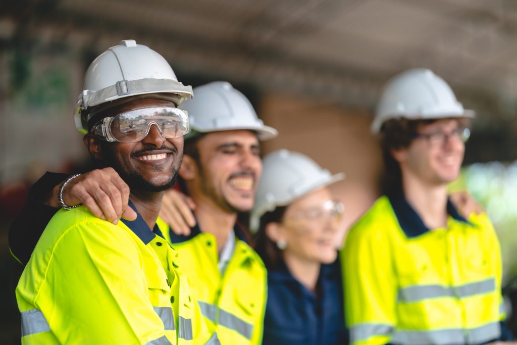 Four construction workers in high-visibility clothing and safety gear stand together smiling, appearing relaxed and happy at the work site.