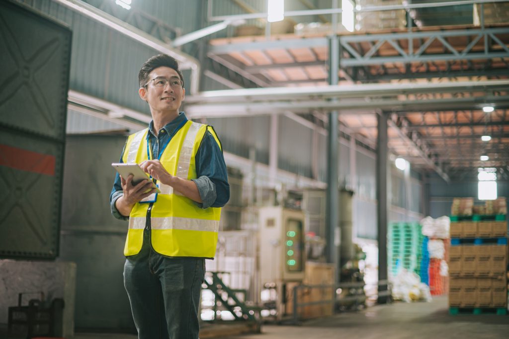 A man wearing a high-visibility vest holds a tablet and looks to the side while standing in a warehouse. Shelves and industrial equipment surround him.