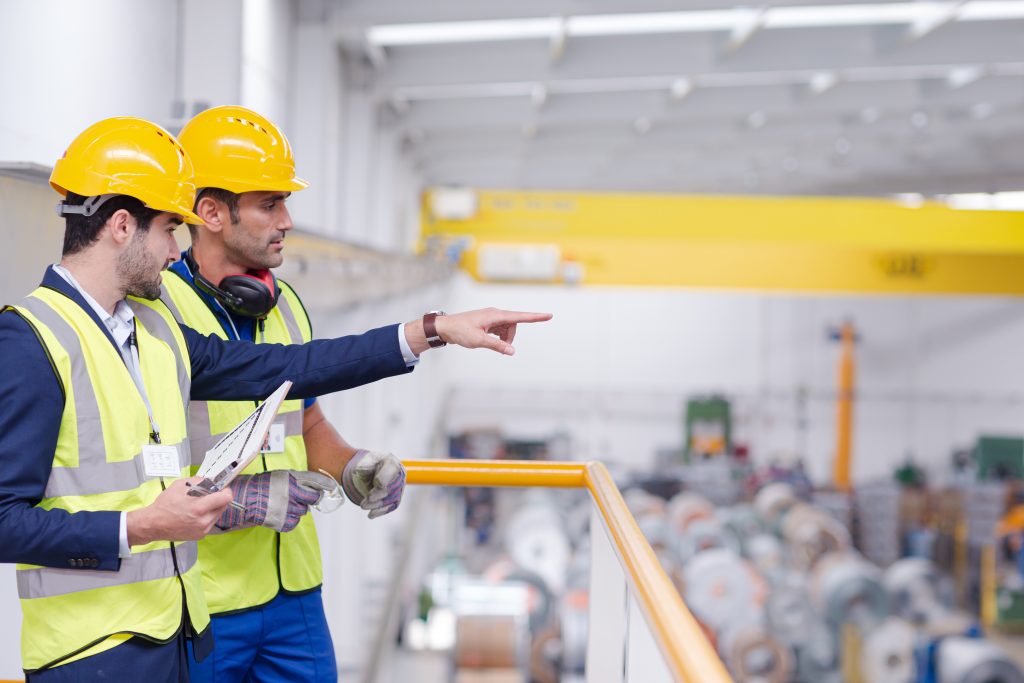 Two workers in safety gear, including hard hats and vests, stand in a factory. One is holding a clipboard, and the other is pointing, both overseeing operations on the factory floor.