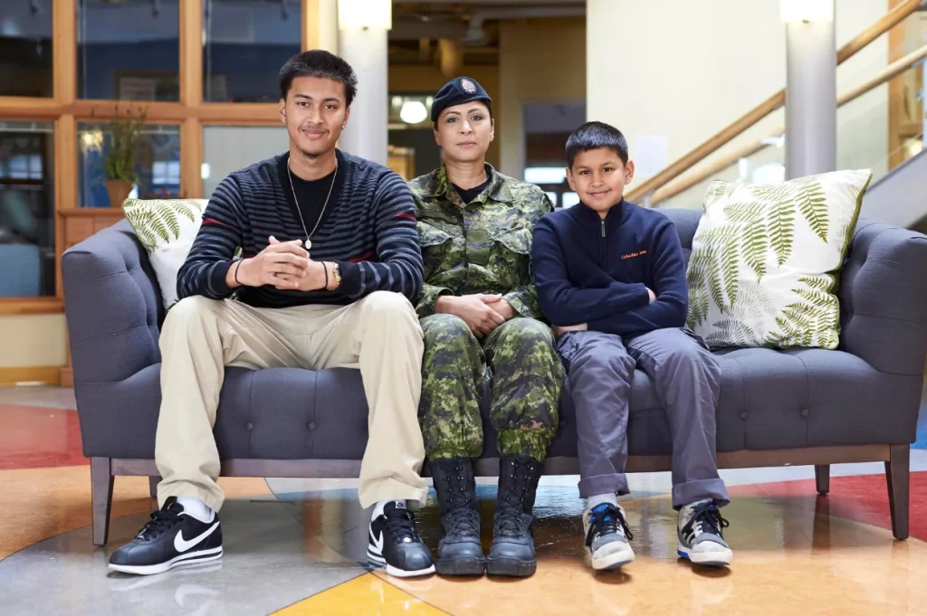Three people sit on a couch. A person in military uniform is seated between two younger people in casual clothing. They are indoors with a staircase in the background.