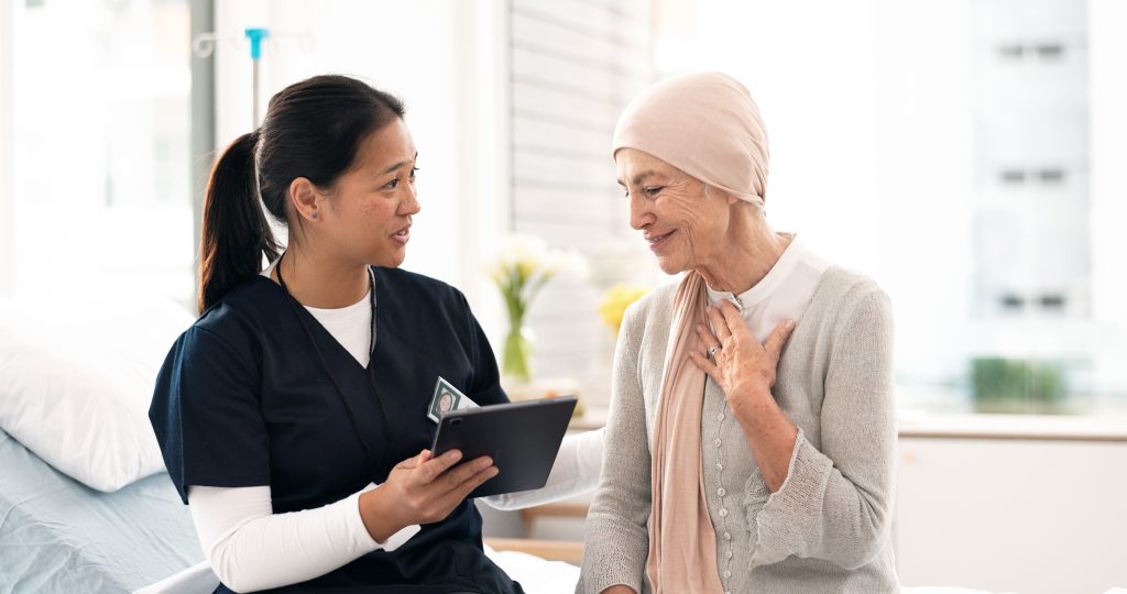 A healthcare professional shows a tablet to an older woman wearing a headscarf, in a hospital room setting.