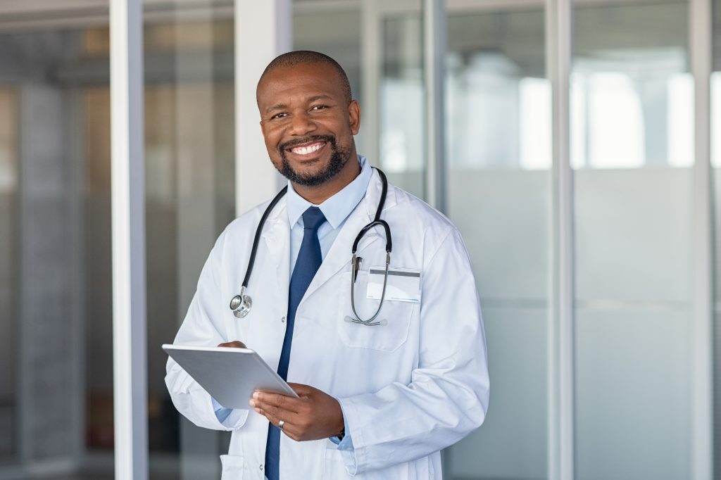 Smiling doctor in a white coat with a stethoscope around his neck holds a tablet, standing in a modern office setting.