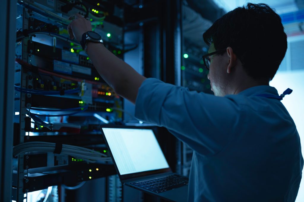Person using a laptop while inspecting and adjusting network equipment in a dimly lit server room filled with cables and blinking lights.