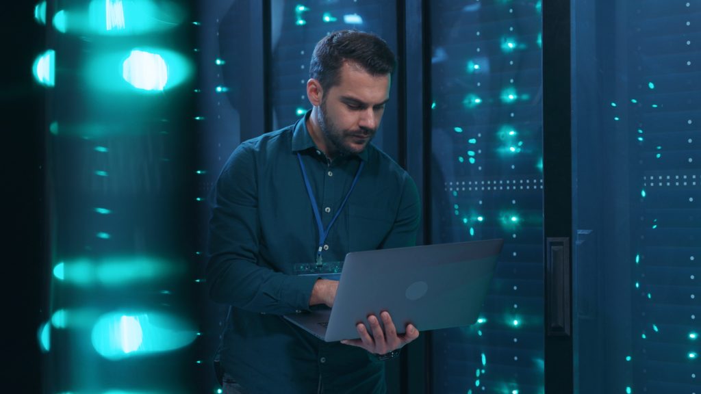 A man stands in a server room holding a laptop, surrounded by illuminated server racks with blue-green lights.
