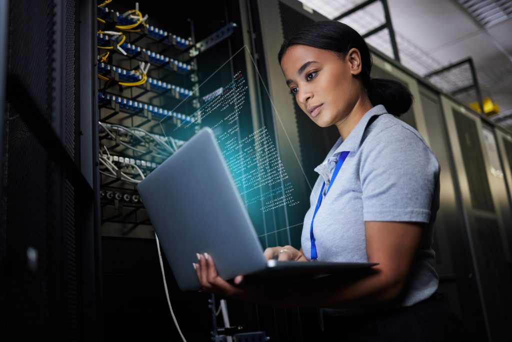 A woman stands in a server room, holding and working on a laptop, with digital code graphics displayed in front of her.