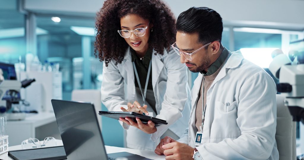 Two scientists in lab coats review information on a digital tablet and laptop in a laboratory setting, with lab equipment visible in the background.