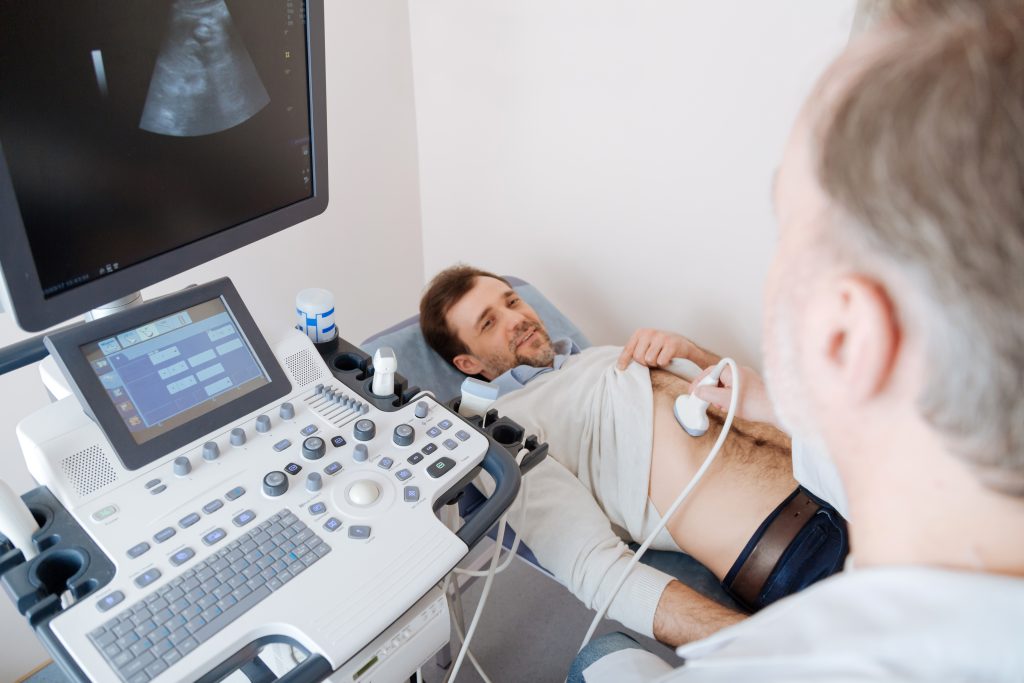 A doctor performs an abdominal ultrasound on a male patient lying on an exam table while monitoring the results on a screen.