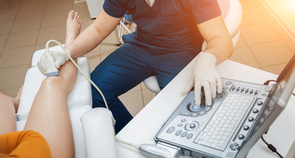 A medical professional performs an ultrasound scan on a patient’s knee, using a handheld device and monitoring the images on a computer.