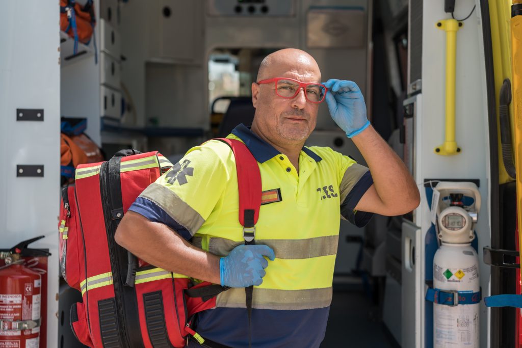 A paramedic wearing a yellow uniform and blue gloves stands in an ambulance, carrying a red medical backpack and adjusting his glasses.