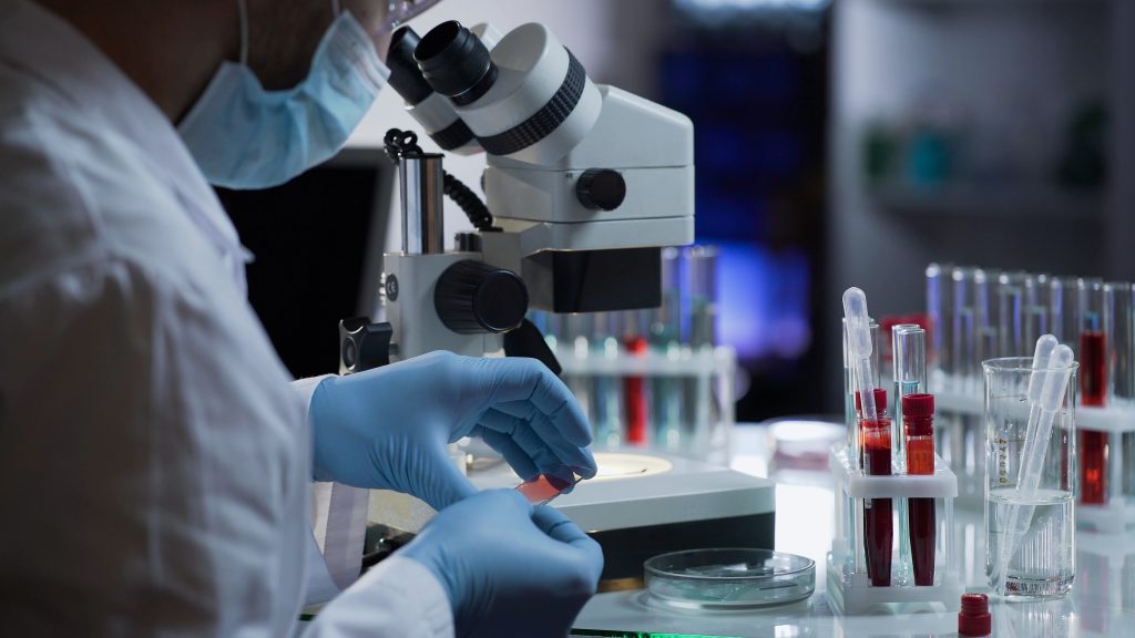 A scientist in gloves and a mask prepares a slide for examination under a microscope, with test tubes containing liquid samples on the lab table.
