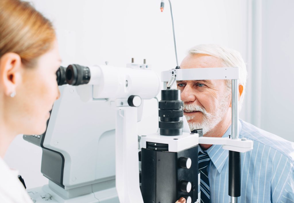 An older man in a blue shirt and tie undergoes an eye examination using a slit lamp; a healthcare professional operates the equipment.
