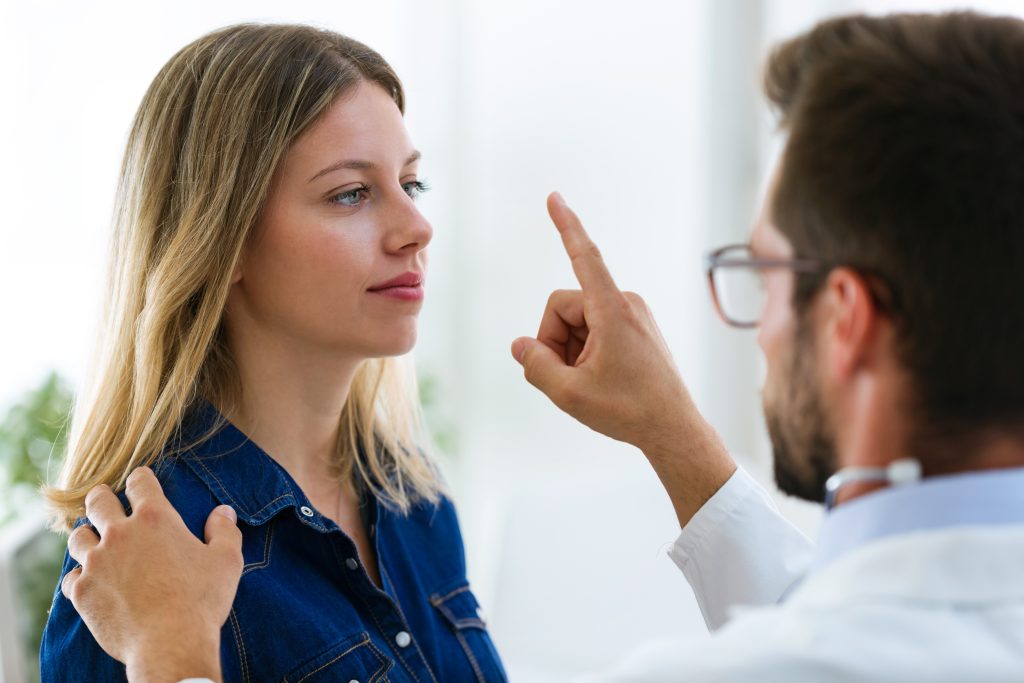 A doctor conducts an eye movement exam, holding a patient’s shoulder and raising a finger in front of her face to track with her eyes.