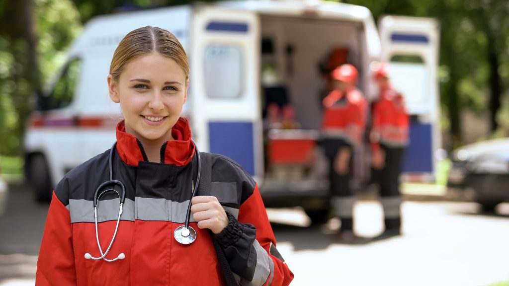 A paramedic in a red uniform with a stethoscope stands in front of an ambulance, with two colleagues and medical equipment visible in the background.
