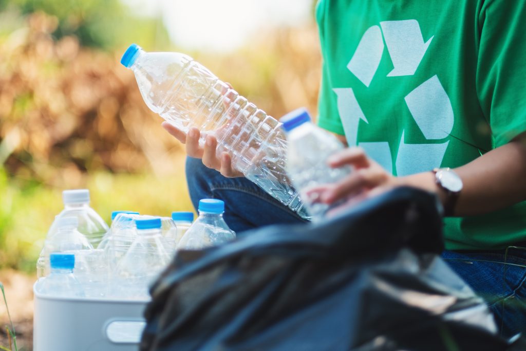 Une personne portant une chemise verte avec un symbole de recyclage ramasse des bouteilles en plastique vides en plein air pour les recycler, en les plaçant dans un sac noir.