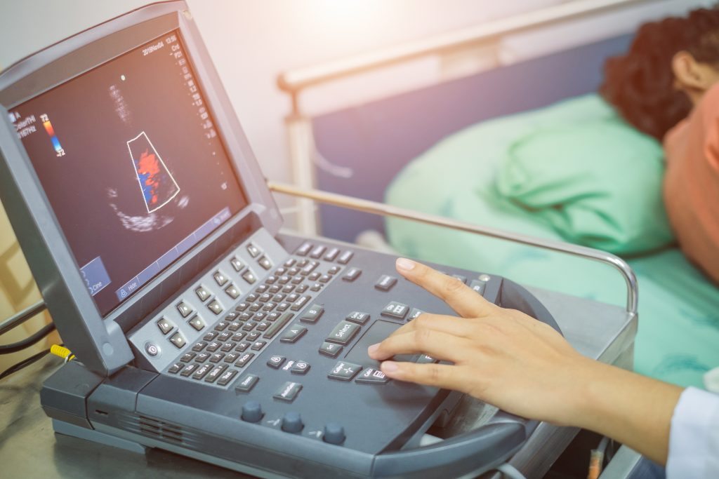 A medical professional operates an ultrasound machine next to a patient lying in a hospital bed.