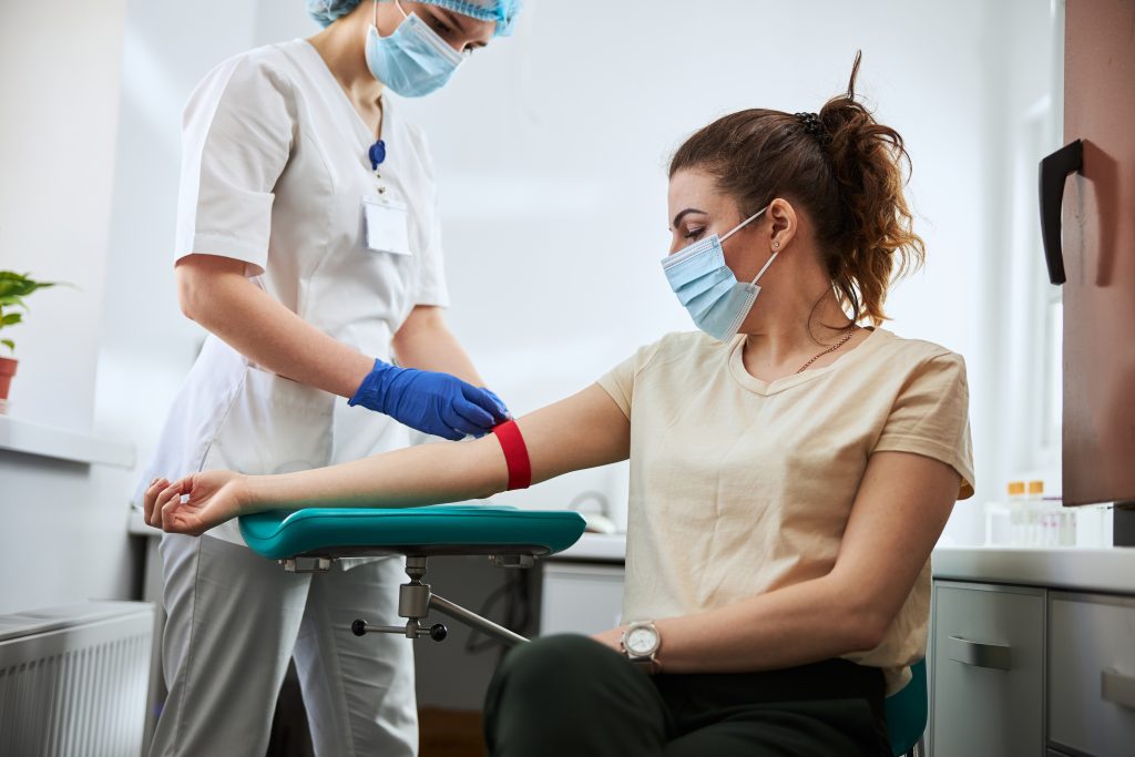 A healthcare worker prepares to draw blood from a seated woman wearing a mask, using a tourniquet on her arm in a medical setting.