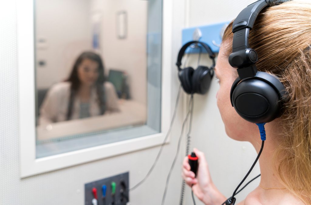 A woman wearing headphones holds a button inside a soundproof booth, while another person observes through a window in a hearing test room.