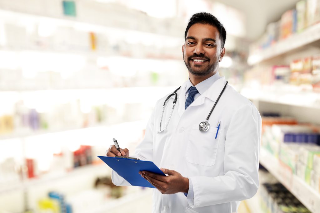 A male doctor in a white coat with a stethoscope stands in a pharmacy, holding a clipboard and pen, smiling at the camera.