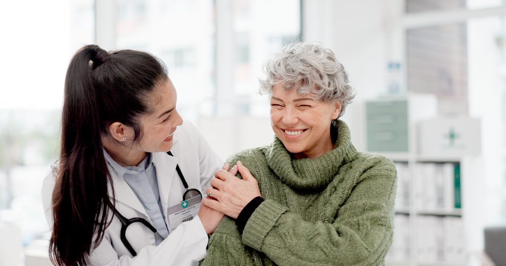 A doctor in a white coat smiles and gently holds the shoulder of an older woman in a green sweater, who is also smiling, in a medical office.