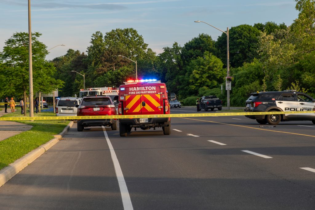 Police and fire vehicles are parked on a road behind yellow caution tape, blocking traffic, with trees and grass visible in the background.