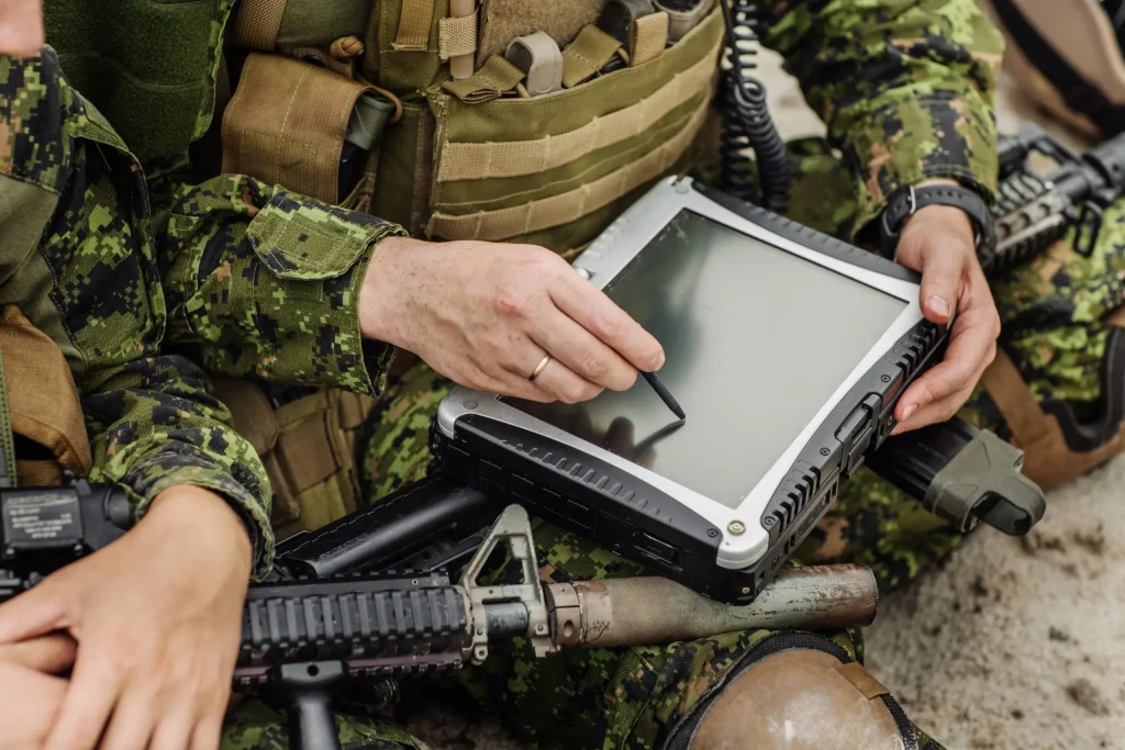 Two soldiers in camouflage uniforms using a rugged tablet while sitting outdoors.