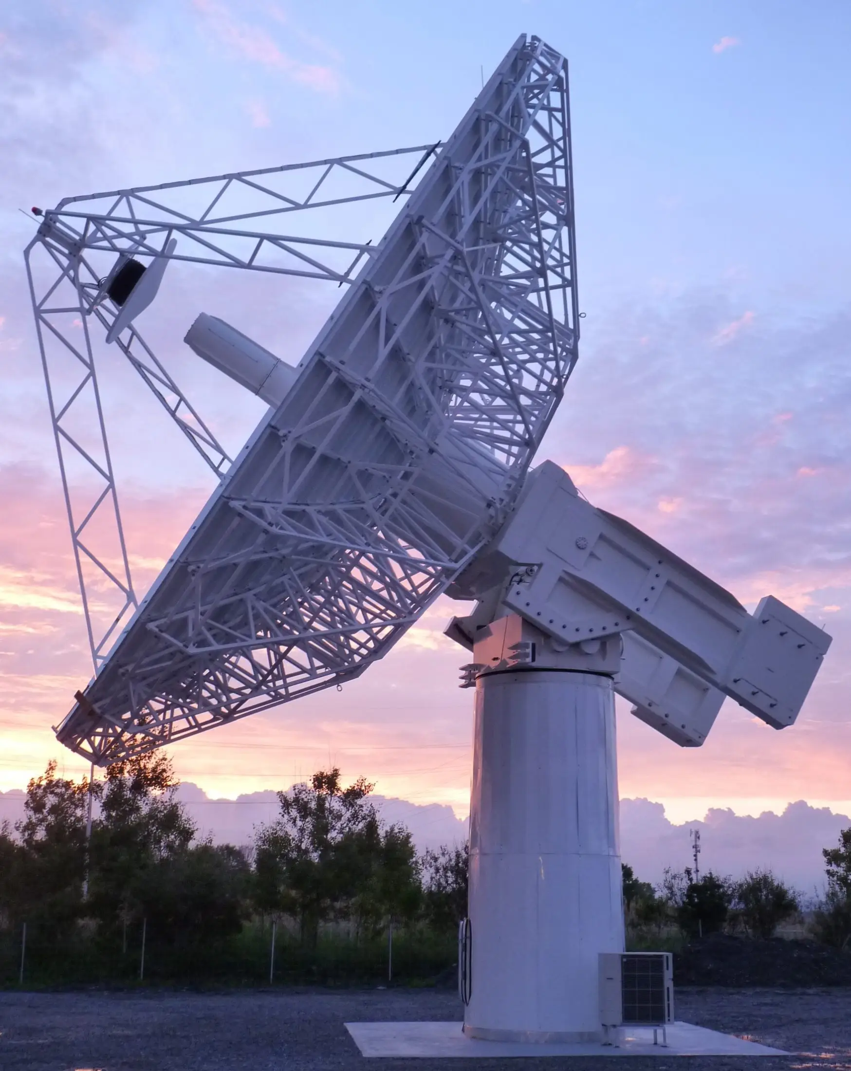 Large radio telescope pointing upward during a sunset with vibrant pink and orange sky in the background.