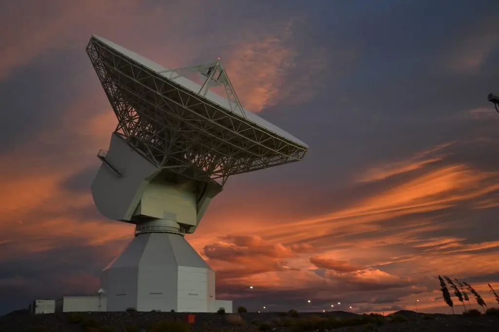 A large radio telescope under a vibrant sunset sky with orange and pink clouds.