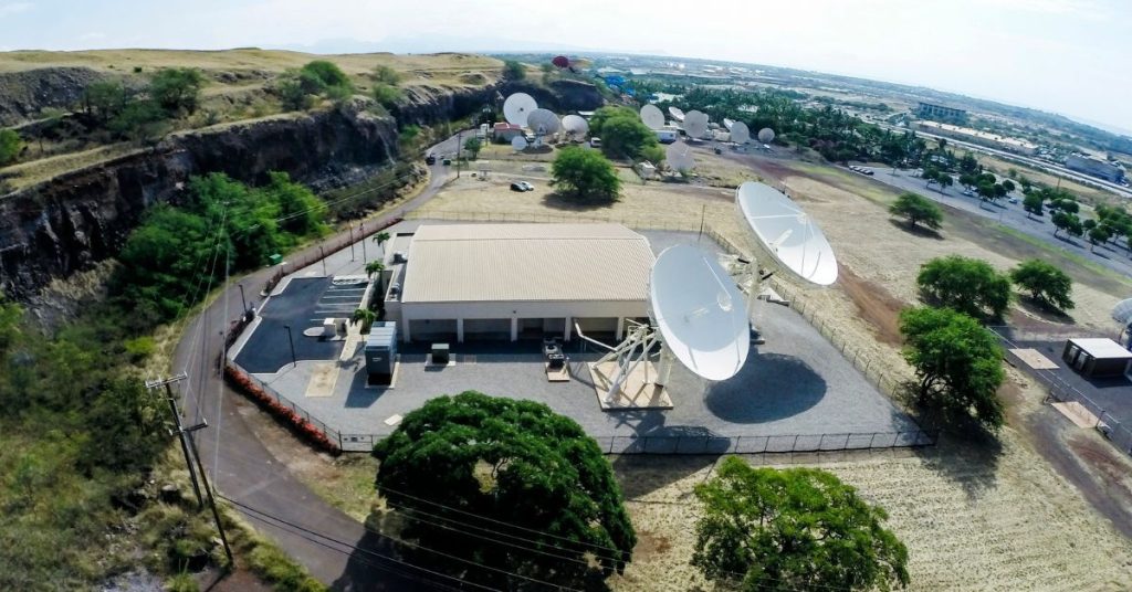 Aerial view of a satellite communication center with numerous large and small dishes, surrounded by greenery and roads, under a clear blue sky.